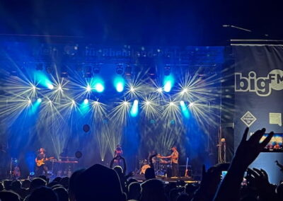 Menschenmenge bei einem Nacht-Konzert, beobachtet eine Band auf der Bühne, umgeben von intensiven blauen und weißen Lichtstrahlen.