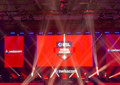 Beleuchtete Bühne mit roten und weißen Lichtern bei einem eSports-Event, Logos von Swisscom und ESL Hero League sind prominent auf großen Bildschirmen zu sehen.