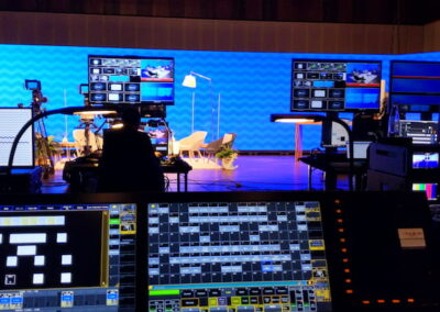 Breiter Blick auf eine Fernsehproduktionsumgebung mit mehreren Bildschirmen und Steuerpulten im Vordergrund. Im Hintergrund ist eine Bühne mit blauem Hintergrund und dekorativen Elementen sichtbar.