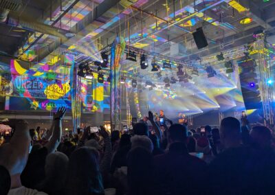 Eine belebte Indoor-Veranstaltung mit vielen Menschen, die einem Konzert beiwohnen. Die Szene ist erleuchtet durch vielfältige, farbenfrohe Lichtstrahlen und große Bildschirme, die lebhafte Grafiken anzeigen.