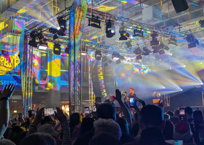 Menschenmenge bei einem Indoor-Konzert, bunte Lichter und Projektionen beleuchten die Bühne, auf der eine Band spielt.