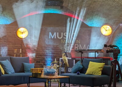 Ein Musikstudio in einem Raum mit Gewölbedecke, beleuchtet mit warmen Lichtern und bunten Lichtstrahlen. Die Einrichtung umfasst ein blaues Sofa mit Kissen, einen Couchtisch, eine Glasvitrine mit Getränken, eine Kamera auf einem Stativ und verschiedene Musikinstrumente. Die Wände sind aus rotem Backstein.