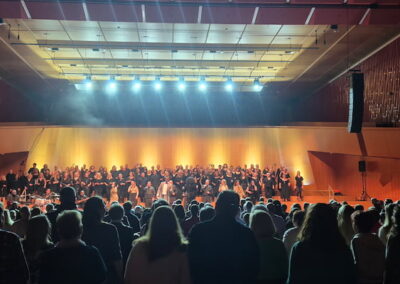 Publikum steht und beobachtet eine große Gruppe von Chorsängern und Musikern, die auf einer beleuchteten Bühne in einem Konzertsaal performen.