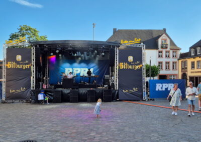 Eine Konzertbühne auf einem belebten Stadtplatz. Menschen verschiedener Altersgruppen gehen umher oder stehen und unterhalten sich. Im Hintergrund sind historische Gebäude sichtbar. Die Bühne zeigt das Logo "RPR1" und ist von Werbebannern der Marke "Bitburger" umgeben. Ein kleines Kind steht alleine vor der Bühne und schaut zu.