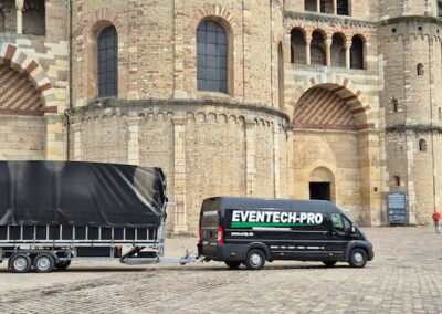 Ein schwarzer Lieferwagen und ein angehängter Anhänger stehen auf einem gepflasterten Platz vor einer beeindruckenden historischen Kirche mit romanischer Architektur. Auf dem Lieferwagen steht "EVENTTECH-PRO".