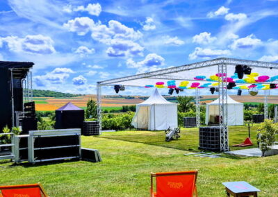 Eine Freiluftbühne auf einer Wiese, ausgestattet mit Lautsprechern, Lichttechnik und bunten Dekorationen, umgeben von Feldern und blauem Himmel.