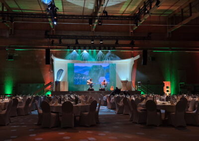 Großer Saal für ein Gala-Dinner vorbereitet mit vielen runden Tischen und weißen Stühlen. Auf der Bühne befinden sich drei Musiker, umgeben von grünen und blauen Lichteffekten.