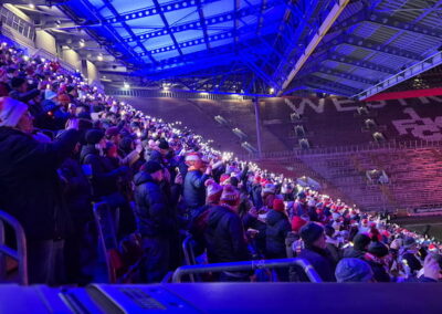Zuschauer, bekleidet mit Wintermützen, stehen und sitzen im beleuchteten Fußballstadion während eines abendlichen Spiels. Die Tribünen sind teilweise gefüllt und in blaues Licht getaucht.