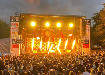 Menschenmenge bei einem Open-Air-Konzert während des Sonnenuntergangs. Die Bühne ist mit intensiven Lichteffekten und Flammen erleuchtet, umgeben von Bäumen.