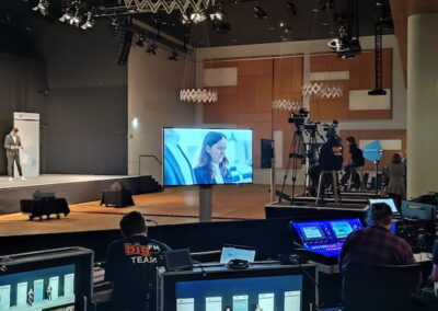 Blick auf einen modernen Konferenzsaal während einer Veranstaltung. Im Vordergrund sind technische Bedienpulte mit Bildschirmen und Technikern. Auf der Bühne stehen zwei Personen, die präsentieren, und ein großer Bildschirm zeigt eine zusätzliche Teilnehmerin in Großaufnahme. Im Hintergrund sind weitere Personen und Kameras zu sehen.
