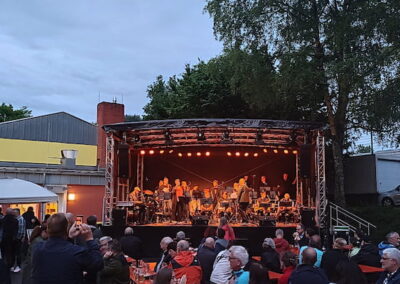 Menschen genießen ein Live-Konzert im Freien bei Abenddämmerung. Die Bühne ist beleuchtet und die Band spielt Musik, während das Publikum auf Stühlen sitzt und steht.