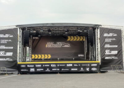 Eine große mobile Bühne mit geöffneten schwarzen Wänden, die Werbung für das ADAC GT Masters Event zeigt. Die Bühne steht auf einem leeren Platz, umgeben von einigen Verkehrsschildern und Bäumen im Hintergrund.