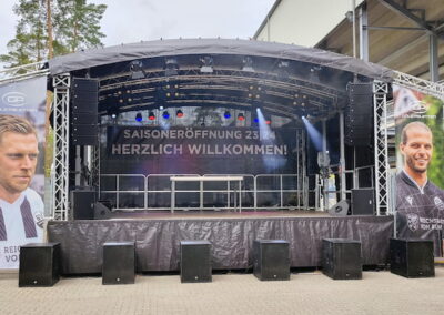 Eine Bühne für eine Saisoneröffnungsveranstaltung im Freien mit zwei großen Bannern, die Fußballspieler zeigen, und einem Willkommensschild "Saisoneröffnung 23, Herzlich Willkommen".