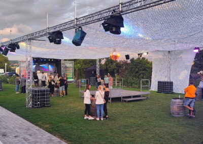 Menschen genießen ein Freiluftkonzert unter einer beleuchteten Bühnenkonstruktion mit Bühnenlichtern und einer Band, die auf einer kleinen Bühne spielt. Die Umgebung ist grün und der Himmel ist bei beginnender Dämmerung leicht bewölkt.