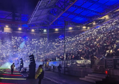 Ein belebtes Stadion bei Nacht, gefüllt mit Menschen, die kleine leuchtende Lichter halten, erzeugt eine funkelnde Atmosphäre unter einem beleuchteten Stahlgerüst.