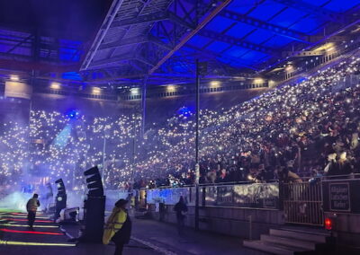 Eine Menschenmenge bei einer nächtlichen Sportveranstaltung in einem offenen Stadion, beleuchtet von vielen kleinen Lichtern, die das Publikum hält, mit blauem Bühnenlicht und Raucheffekten im Vordergrund.