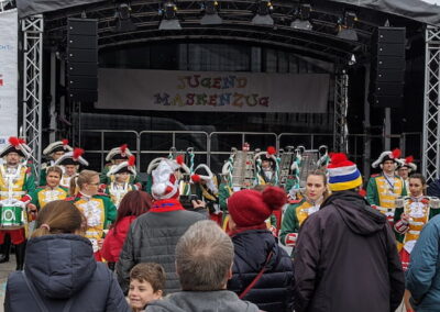 Eine Gruppe junger Menschen in traditionellen Karnevalskostümen mit Trommeln und anderen Musikinstrumenten steht auf einem öffentlichen Platz vor einer Bühne, auf der "Jugend Maskenzug" steht. Zahlreiche Zuschauer, darunter Kinder und Erwachsene, beobachten die Darbietung.