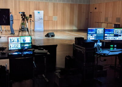 Ein dunkler Raum mit technischer Ausrüstung für eine Medienproduktion, darunter mehrere Monitore, Kameras und Lichttechnik, in einem Saal mit Holzboden und -wänden. Eine Person bedient die Ausrüstung.