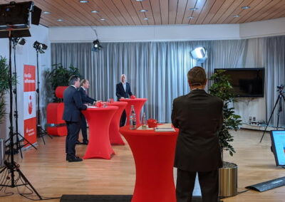 Innenraum einer Veranstaltungshalle mit drei Männern und mehreren roten Stehtischen. Zwei Männer unterhalten sich an einem Tisch, während ein dritter Mann sie filmt. Im Hintergrund sind große Bildschirme und Kameraausrüstung zu sehen.