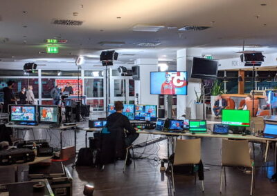 Innenraum eines Stadions umgebaut für Medienproduktion. Zahlreiche Bildschirme, Kameras und Arbeitsstationen sind sichtbar. Im Hintergrund sind Personen und Werbebanner erkennbar.