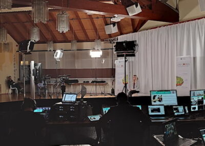 Innenansicht eines Veranstaltungssaals mit technischem Equipment und mehreren Personen, die an Computern arbeiten. Im Hintergrund sind Kameras und eine beleuchtete Bühne zu sehen.