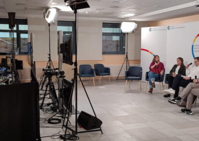 Fünf Personen sitzen in einer Reihe auf Stühlen in einem hell beleuchteten Raum und nehmen an einer Podiumsdiskussion teil. Im Vordergrund sind technische Ausrüstungen und Kamerateams zu sehen, die die Diskussion aufzeichnen.