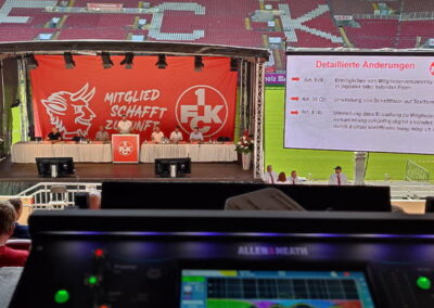 Blick von einem technischen Mischpult auf eine Mitgliederversammlung im Fußballstadion. Die Bühne ist mit einem großen Banner mit der Aufschrift "MITGLIED SCHAFFT ZUKUNFT" und einem Stier-Logo dekoriert. Einige Personen sitzen in den vorderen Reihen und schauen auf eine Leinwand, die detaillierte Änderungen zeigt.