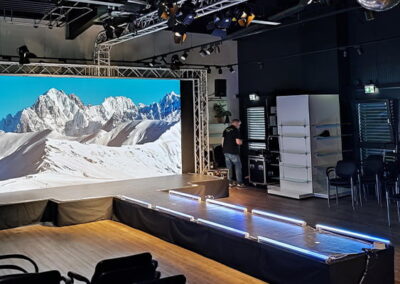 Innenansicht eines Veranstaltungsraumes mit Stühlen, einer Bühne und einem großen Bildschirm, der ein Bergpanorama zeigt. Beleuchtungseinrichtungen hängen von der Decke und eine Person steht am Rand des Raumes.