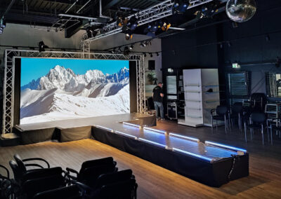 Innenansicht eines Veranstaltungsraumes mit einer großen Bühne, auf der ein Bildschirm ein Bergpanorama zeigt. Im Raum sind Stühle, technische Ausrüstung und eine Person, die Ausrüstung justiert.