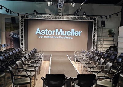 Ein leerer Vortragssaal mit mehreren Reihen schwarzer Stühle, gerichtet auf eine große Leinwand, die das Logo und den Slogan "AstorMueller - Tech meets Shoe Excellence." zeigt.