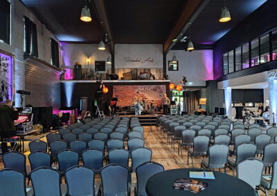 Innenansicht eines Konzertsaaals mit blauen Stühlen, die auf eine Bühne ausgerichtet sind, wo Musiker ihre Instrumente einrichten. Der Raum ist beleuchtet und dekoriert mit musikbezogenen Bannern.