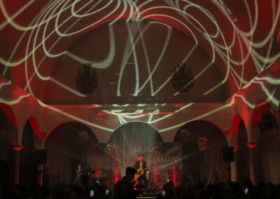Eine Band spielt auf einer Bühne in einem beleuchteten Saal mit roten und weißen Lichtmustern an der Decke während eines Events namens "Music in Germany".