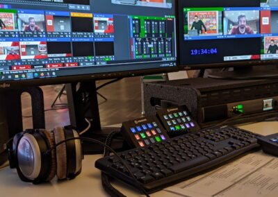 Ein Arbeitsplatz in einer Fernsehregie mit mehreren Monitoren, die verschiedene Live-Feed-Bilder zeigen. Auf dem Tisch befinden sich eine Tastatur, eine Maus, ein Mischpult und Kopfhörer. Eine Hand bedient die Maus.