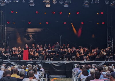 Ein großes Orchester und eine Sängerin in rotem Kleid treten auf einer Freiluftbühne vor einem Publikum bei Tageslicht auf. Die Bühne ist mit roten Lichtern beleuchtet und das Publikum sitzt dicht gedrängt, umgeben von Bäumen.