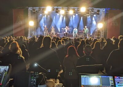 Menschenmenge von hinten betrachtet bei einem Nacht-Konzert. Im Vordergrund sind Bedienpulte für die Licht- und Tonsteuerung zu sehen, die Bühne wird durch helle Scheinwerfer erleuchtet.