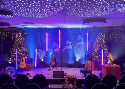 Eine festlich dekorierte Bühne mit Weihnachtsbaum, Geschenken und beleuchteten Rentier-Silhouetten. Der Raum ist mit blauen und violetten Lichtern beleuchtet und Stühle sind vor der Bühne angeordnet.
