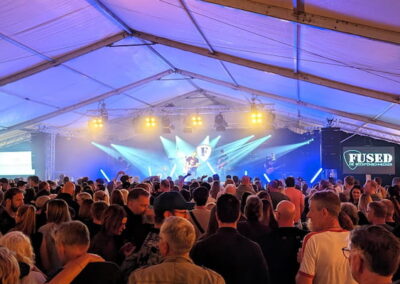 Menschenmenge genießt ein Live-Konzert in einem großen, beleuchteten Zelt. Auf der Bühne sorgen blaue und weiße Lichter für eine dynamische Atmosphäre.