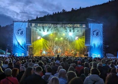 Menschenmenge bei einem Open-Air Konzert in einem Bergtal bei Dämmerung. Auf der Bühne performt eine Band, umgeben von Bäumen und Hügeln.