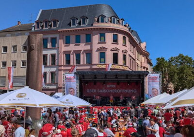 Menschenmenge in roten Outfits auf einem belebten Stadtplatz, der von historischen Gebäuden umgeben ist, während einer Saisoneröffnungsveranstaltung mit einer Bühne im Hintergrund.