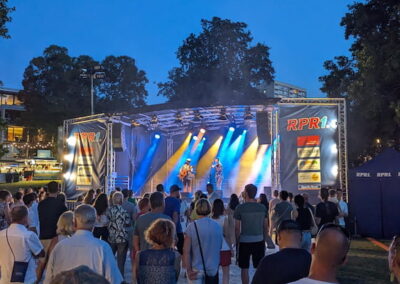 Menschenmenge steht abends vor einer beleuchteten Bühne im Freien, auf der eine Band spielt. Die Atmosphäre ist von Bühnenlichtern und Dämmerung geprägt.