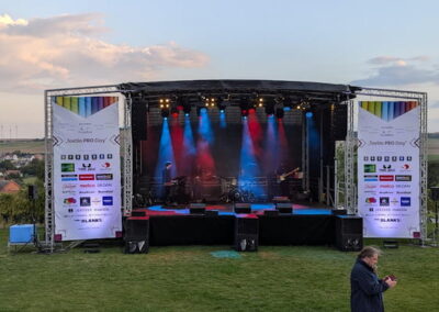 Eine große Open-Air Bühne mit leuchtenden Scheinwerfern und bunten Lichtern bei einem Musikfestival. Rechts und links der Bühne sind große Banner mit Sponsorenlogos. Im Vordergrund steht eine Person, die auf ihr Handy schaut. Im Hintergrund ist eine ländliche Landschaft sichtbar.
