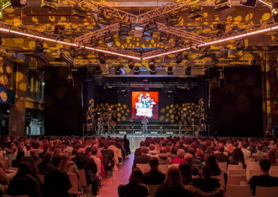 Innenansicht einer großen Eventhalle mit zahlreichen Zuschauern, die auf Stühlen sitzen und auf eine Bühne mit einer großen Leinwand schauen, auf der eine Präsentation läuft. Die Halle ist mit farbigen Lichtern und moderner Architektur ausgestattet.