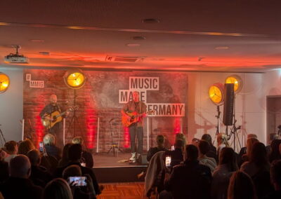 Zwei Musiker spielen Gitarre auf einer Bühne mit der Aufschrift "Music Made in Germany". Sie werden von Scheinwerfern beleuchtet, vor einem Publikum, das teilweise Fotos macht. Die Atmosphäre ist warm und einladend.