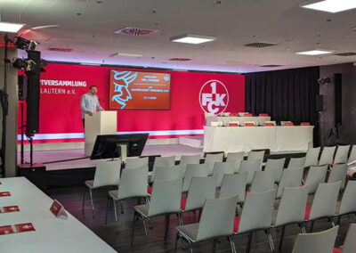 Ein Konferenzraum mit roter und weißer Dekoration, vorbereitet für eine Mitgliederversammlung des 1. FC Kaiserslautern. Auf der Bühne ist ein Rednerpult und ein großes Banner mit dem Vereinslogo. Reihen von leeren weißen Stühlen sind auf das Podium ausgerichtet, und einige Personen sind mit den letzten Vorbereitungen beschäftigt.