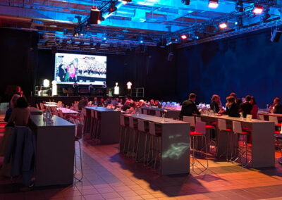 Eine Veranstaltungshalle bei Nacht, beleuchtet in rotem und blauem Licht. Personen sitzen an Tischen und schauen auf einen großen Bildschirm, auf dem eine Präsentation läuft.